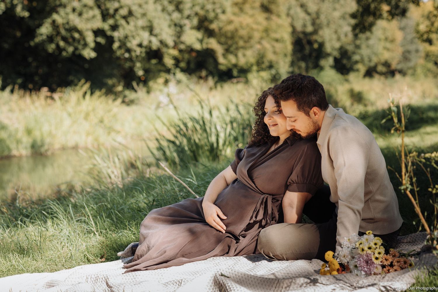 Expectant couple sitting on a picnic blanket by a peaceful lake, the man embracing his pregnant partner, with wildflowers beside them.