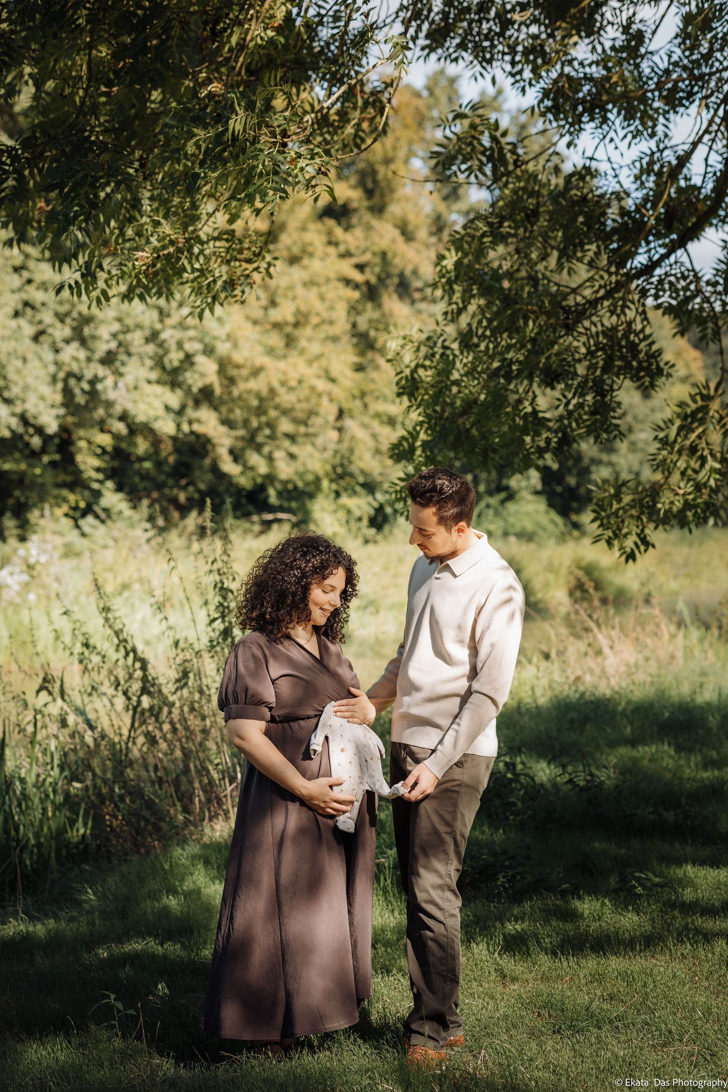 Expecting couple in a sunlit green park holding a small baby garment together during an outdoor maternity photoshoot under leafy trees.