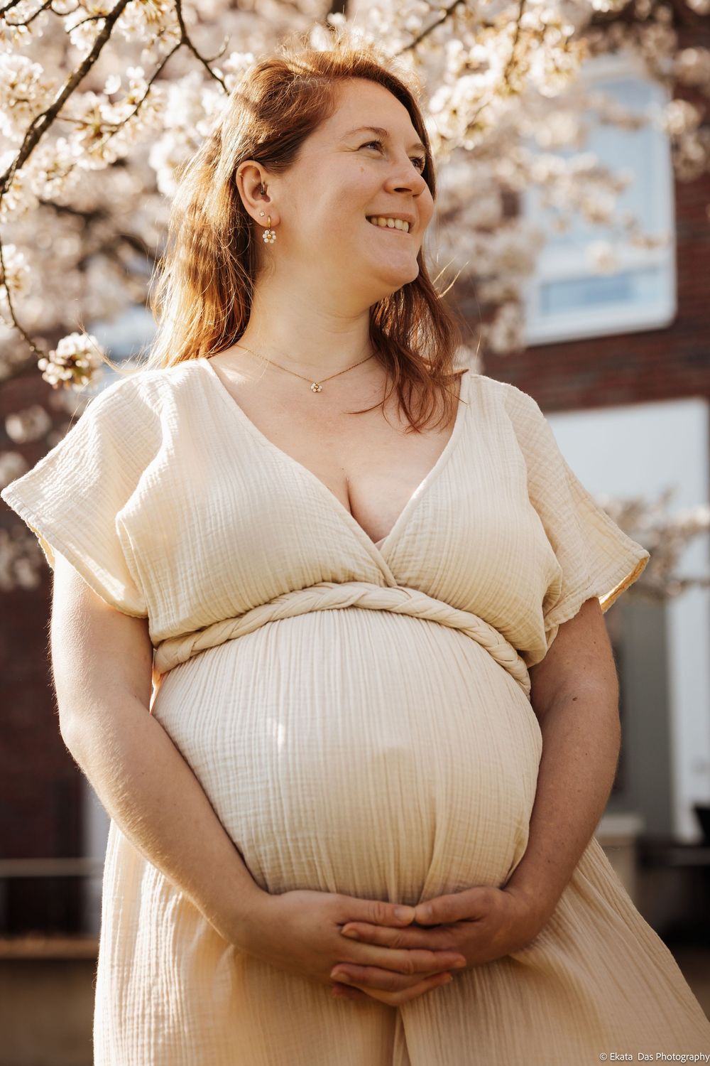 Woman in cream dress poses joyfully among blooming cherry blossoms on a sunny spring day.