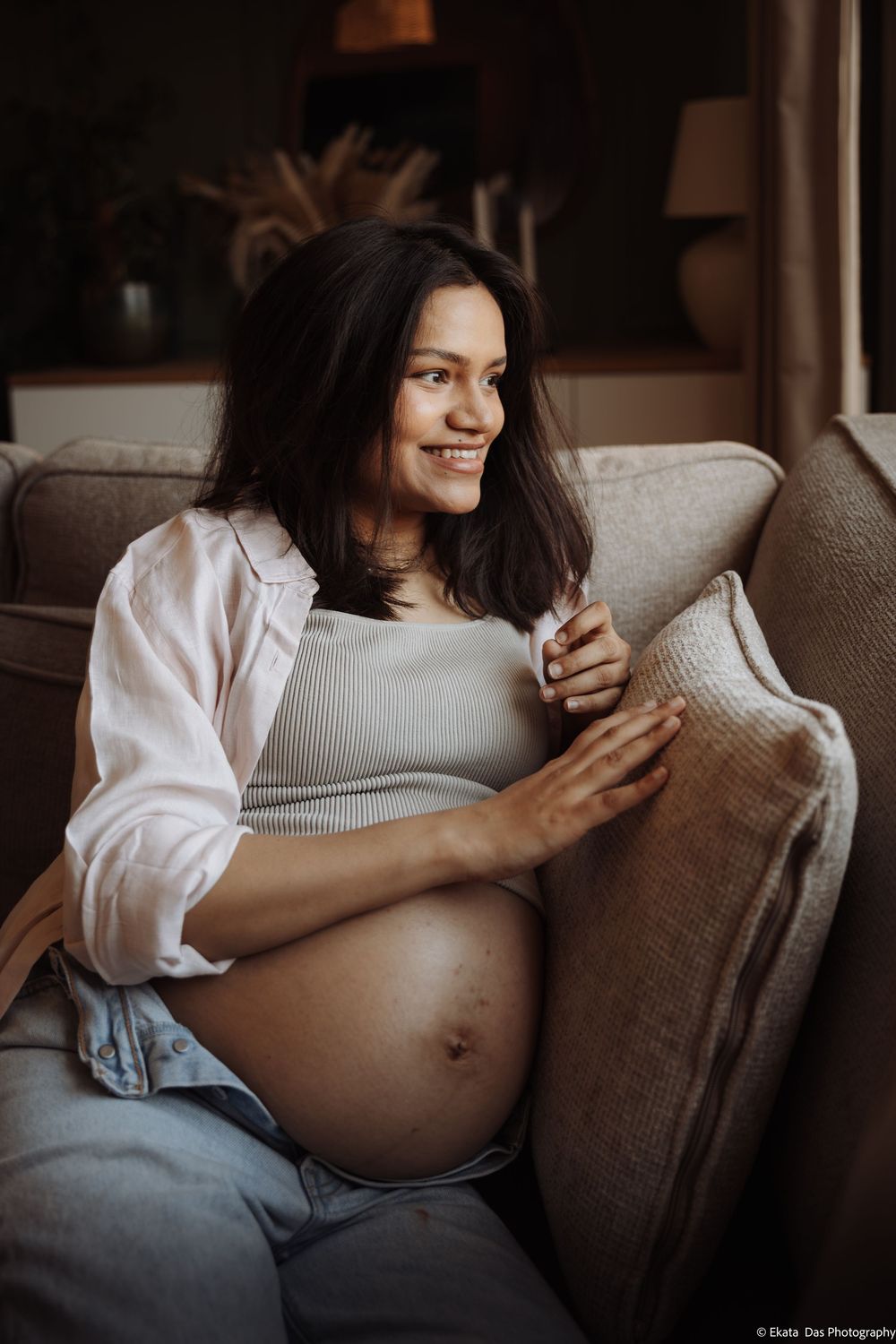 Smiling pregnant woman sitting on a couch, cradling her baby bump in a cozy home setting.