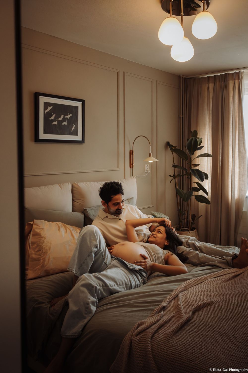 Couple relaxing on bed, pregnant woman lying on man's lap in cozy bedroom with warm lighting and plants.