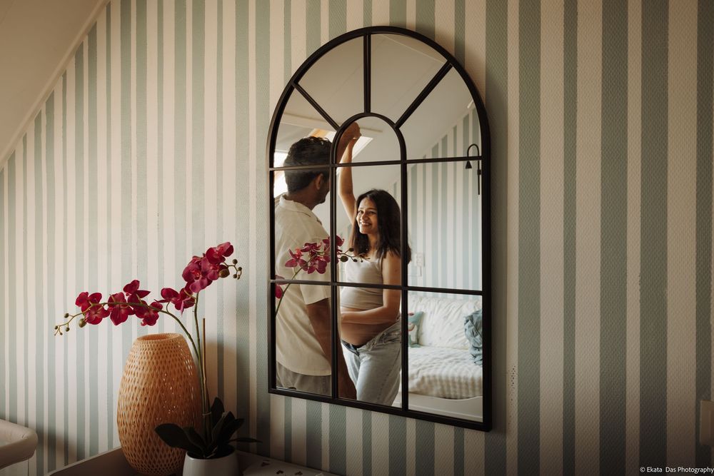 Pregnant woman and partner sharing a tender moment reflected in an arched window mirror in a striped room.