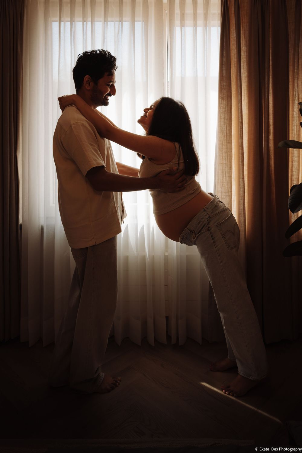 Pregnant couple sharing a tender moment by curtained window in warm backlit silhouette.