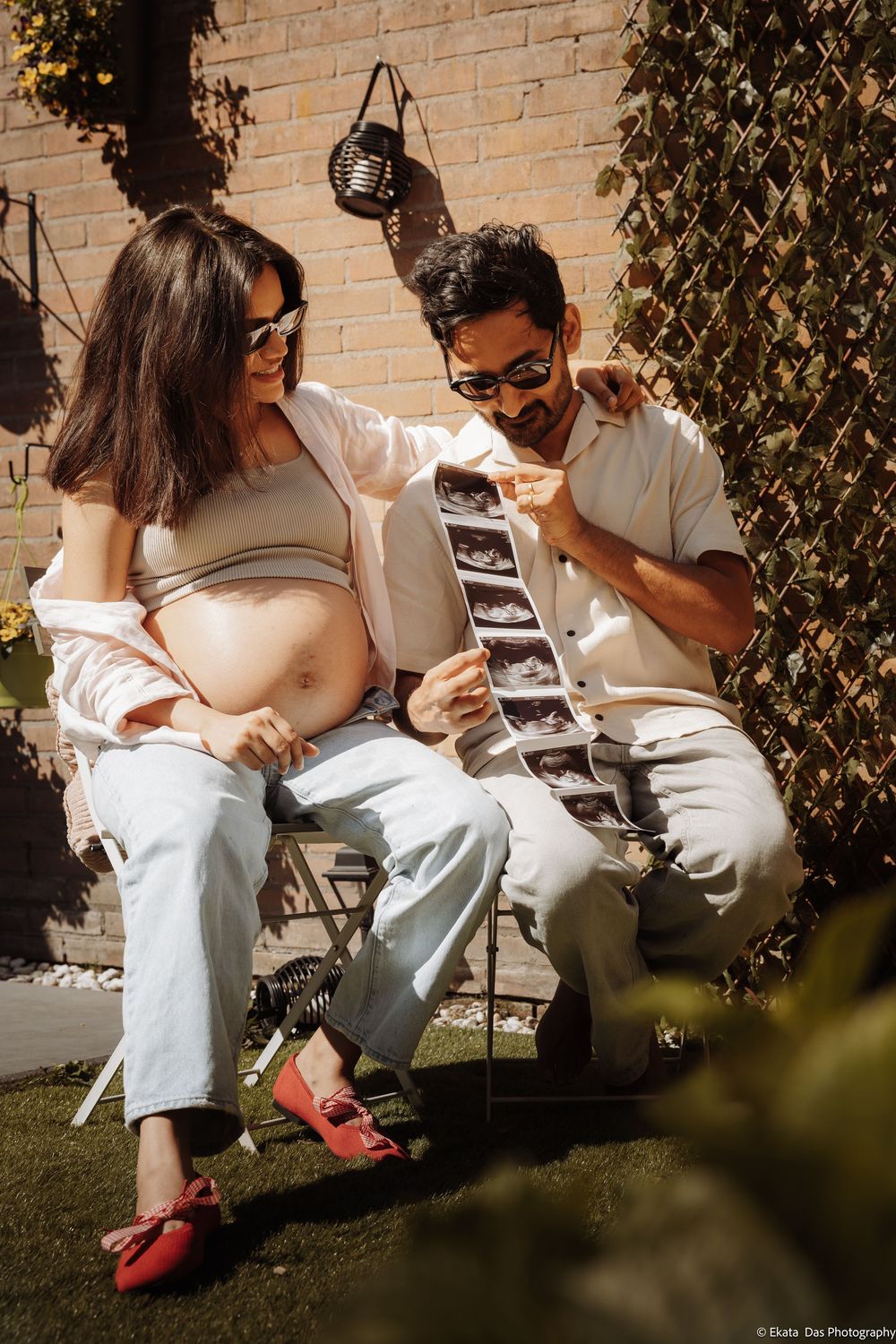 Pregnant couple sitting outdoors in sunlight, examining ultrasound scans together, woman showing baby bump.