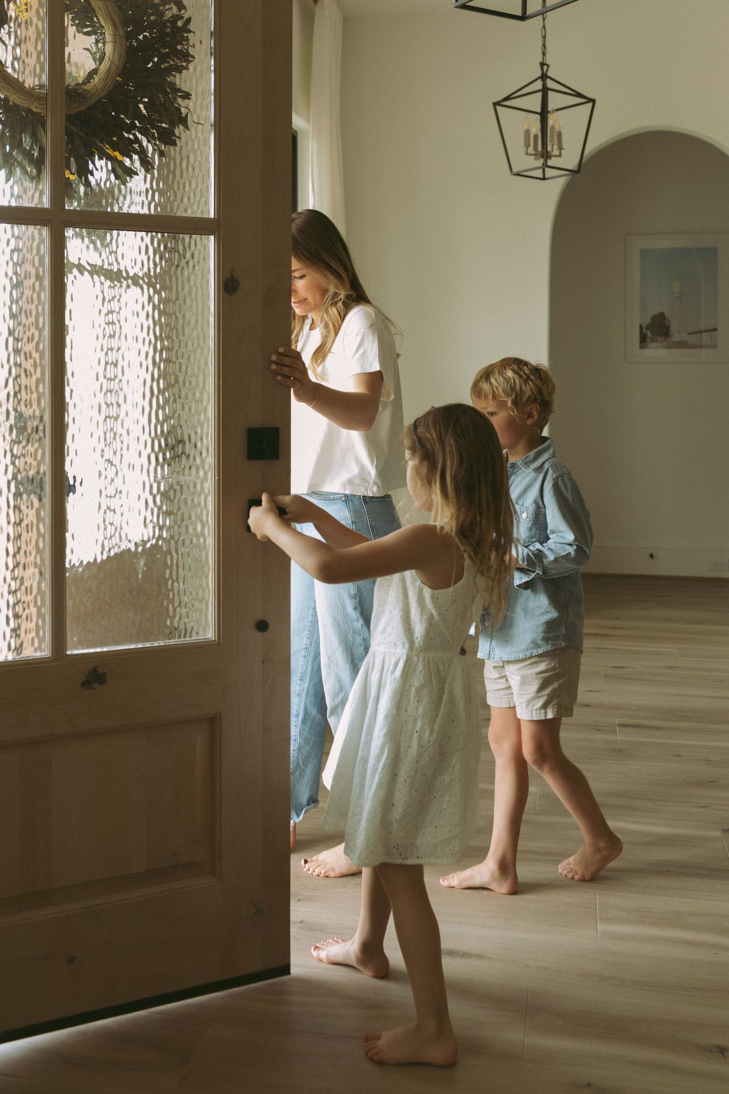 Three people stand barefoot by a glass door in a bright room with hardwood floors and lantern pendant lighting.