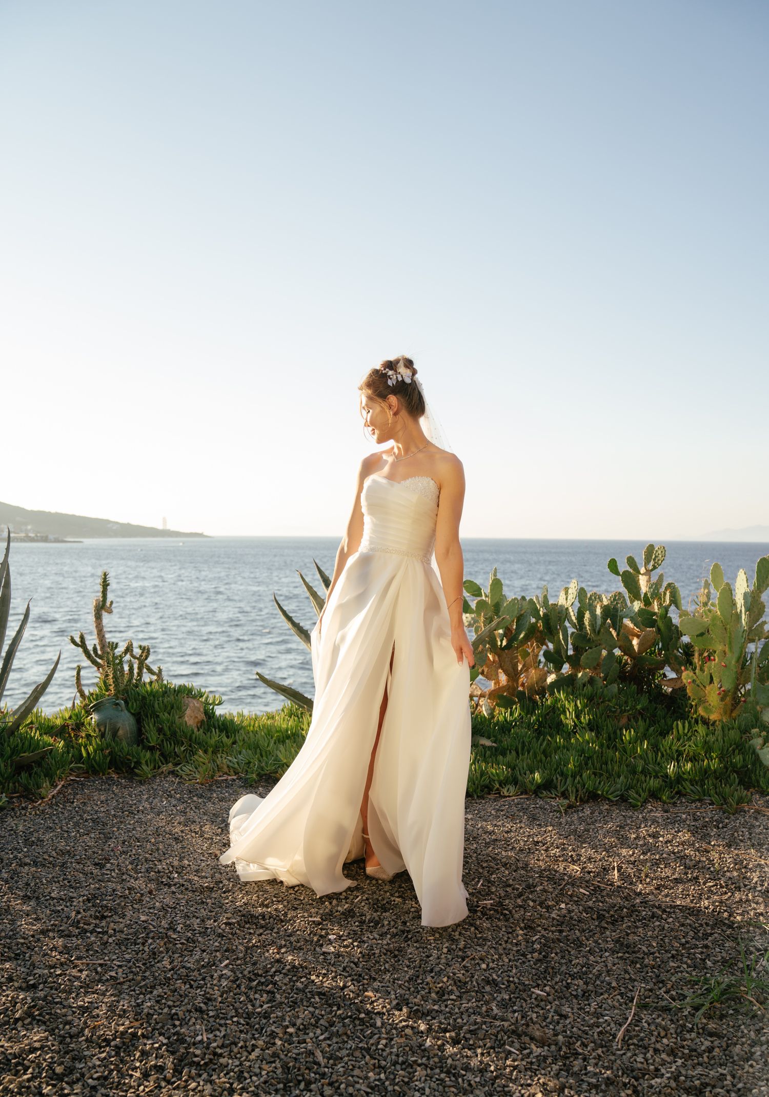 A flowing white wedding dress is showcased against a coastal backdrop with ocean views and native plants at sunset.