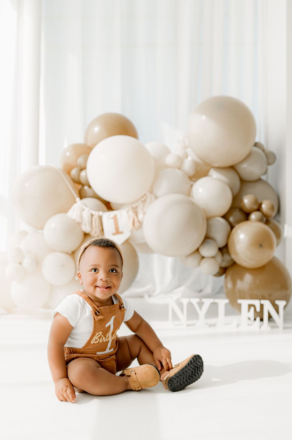 Baby in brown overalls sits among cream and beige balloon decorations during a milestone photoshoot.