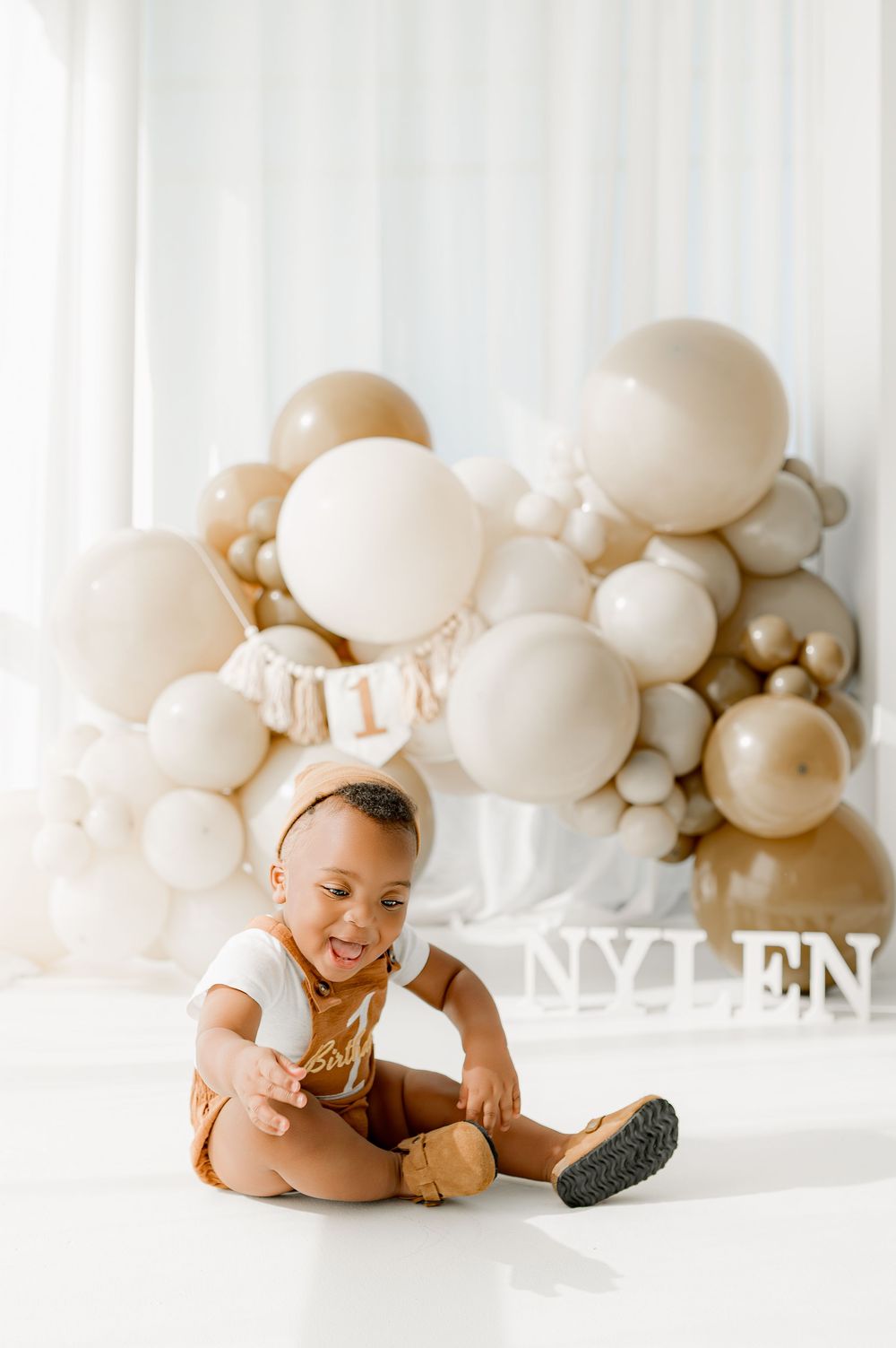 A baby lies on a white floor with an elegant display of neutral-colored balloons creating a dreamy backdrop.