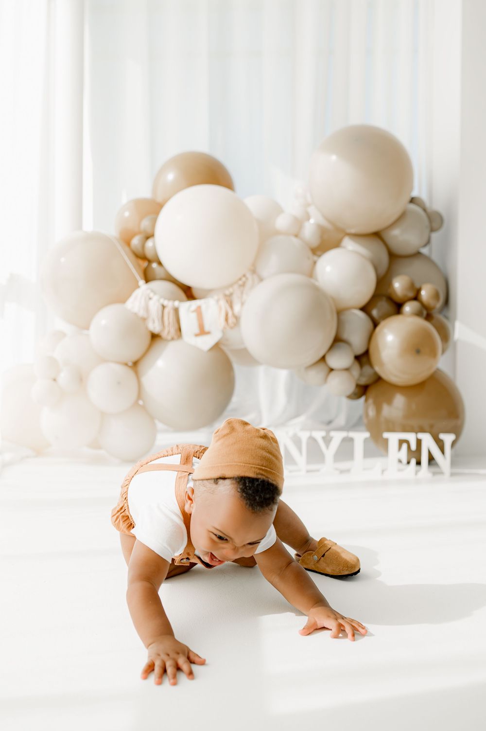 Baby in brown hat crawls on white surface with elegant beige and gold balloon arch display in background.