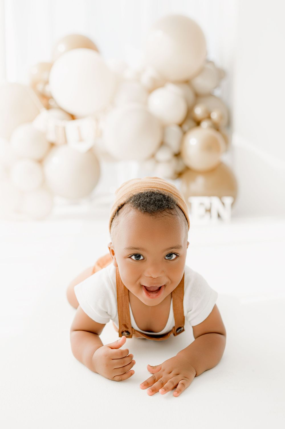 Baby in white onesie smiling and lying on white background with soft white balloons in background.