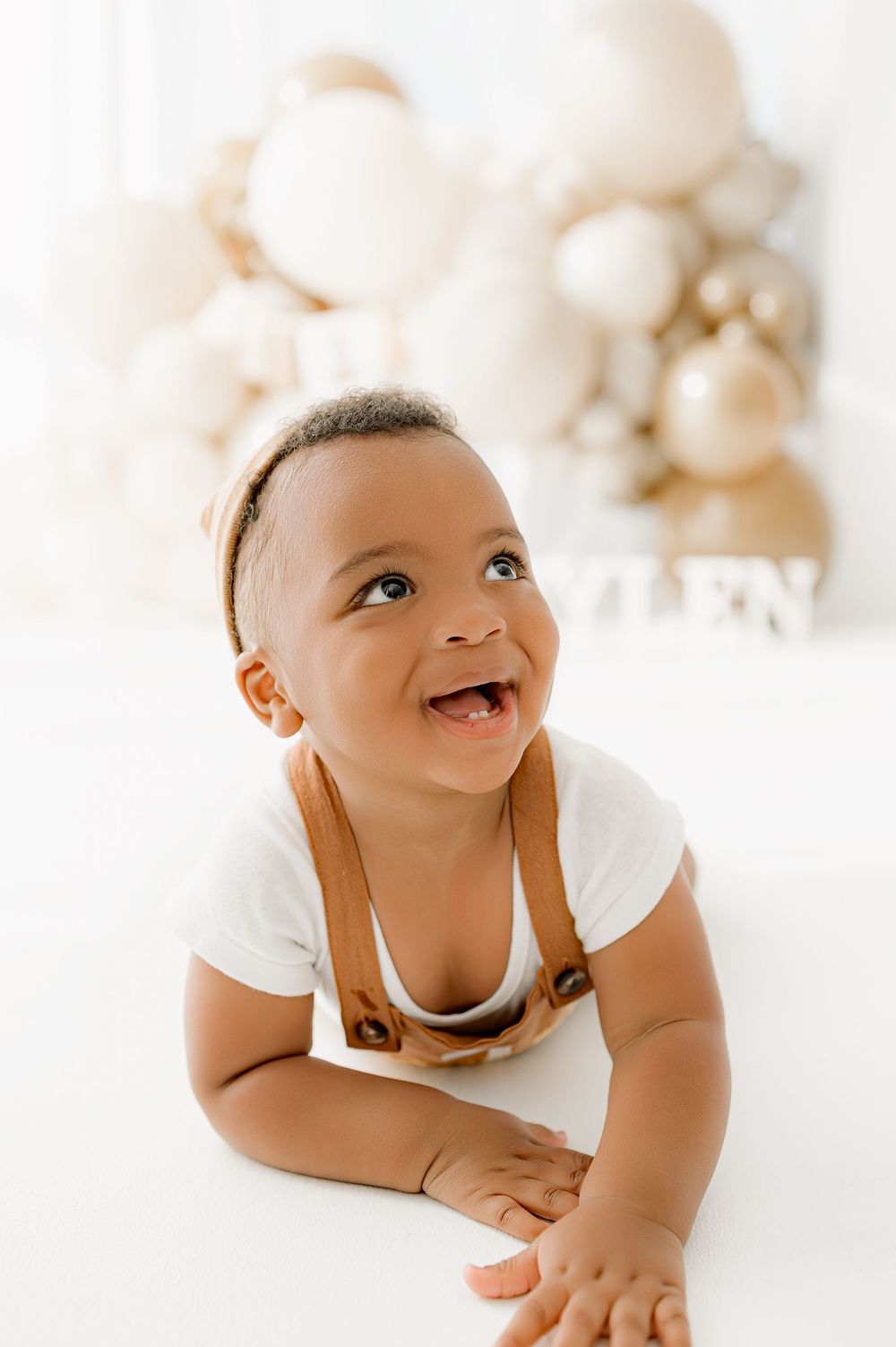 Joyful baby in white onesie lying on stomach against soft white background with blurred decorative elements.