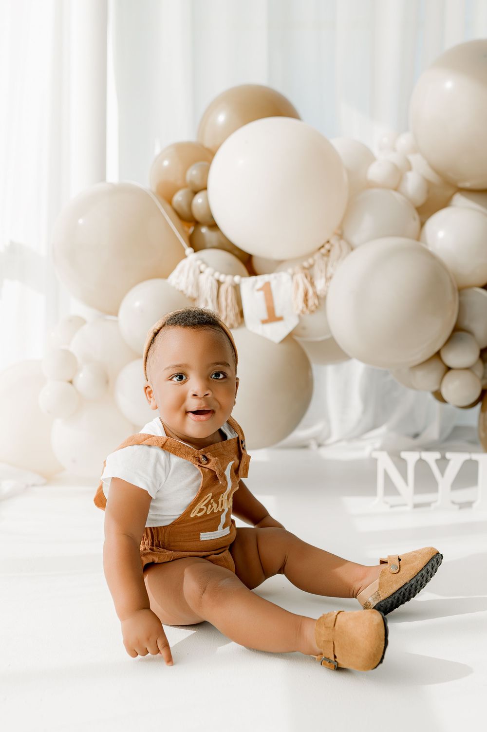Baby in brown outfit poses against white balloons during milestone photo session.