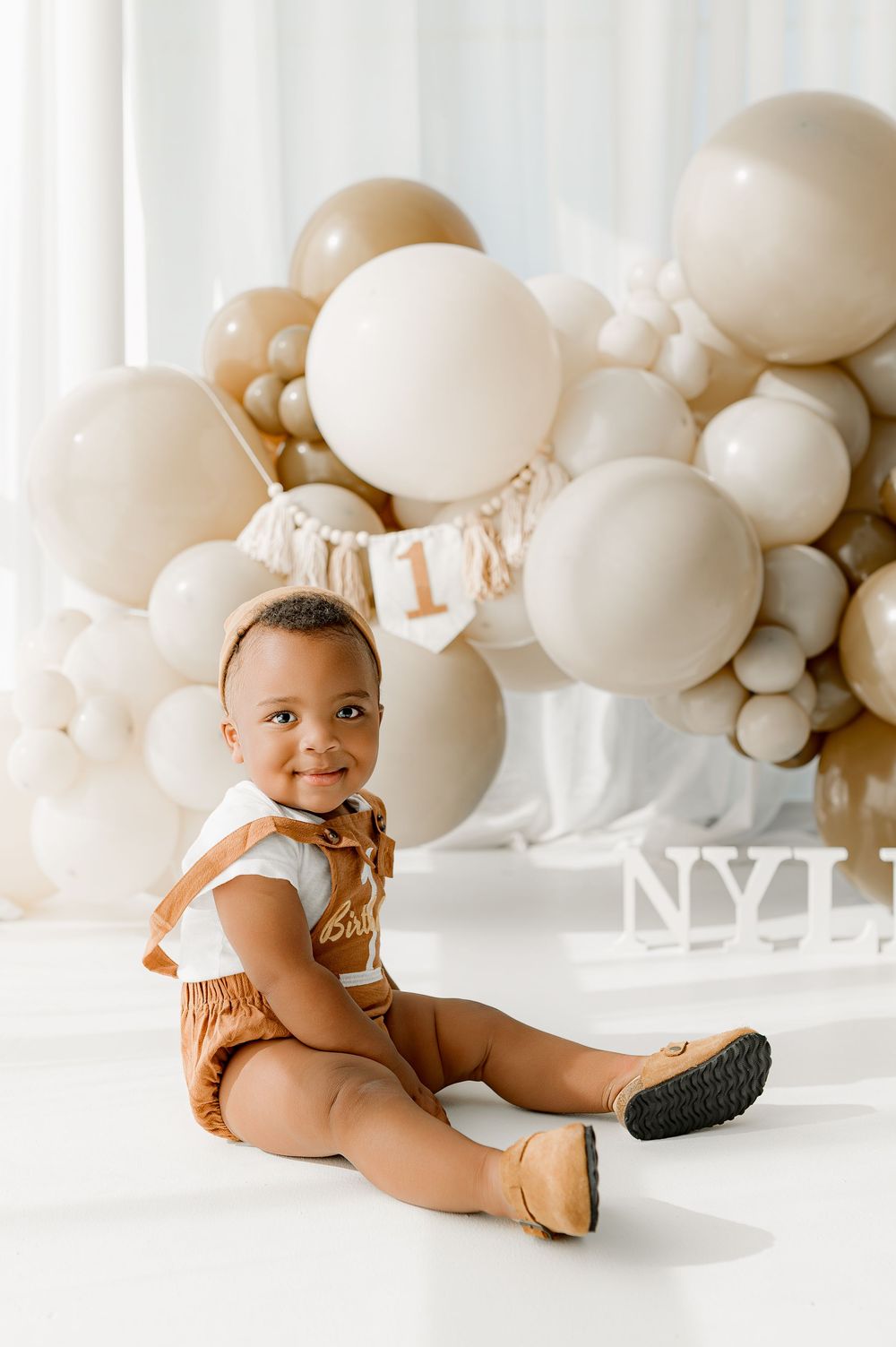 Baby in white top and brown shorts sits on floor with white balloon arch backdrop in minimalist studio setting.
