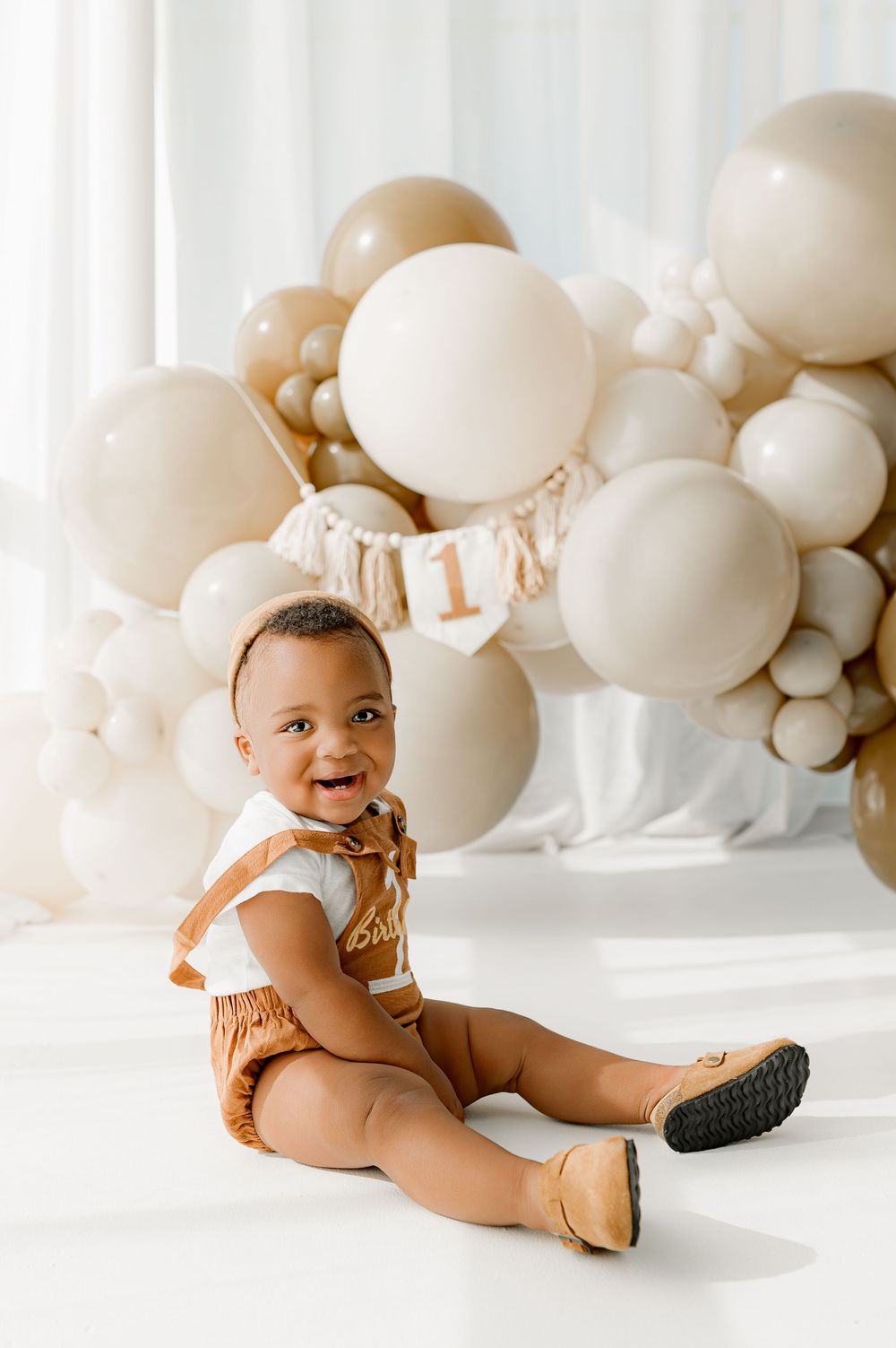 Baby sits on white floor with neutral beige balloon arch in background, creating minimalist cake smash photo setup.