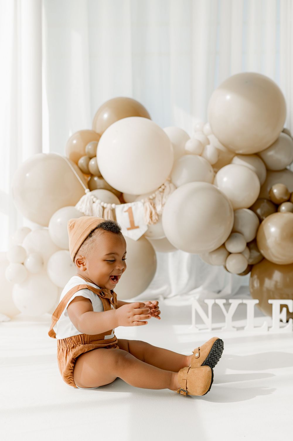 Baby in brown outfit lies on white floor surrounded by cream and beige balloons during photo session.