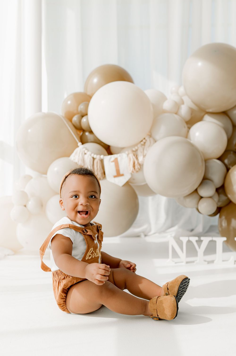 A baby in a brown romper sits on the floor surrounded by white and beige balloons for a photoshoot.