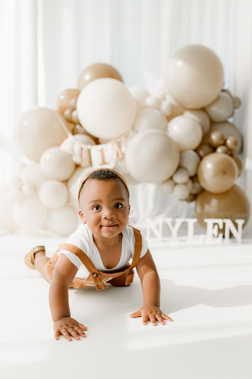 A baby in a white tank top crawls on a white surface with cream-colored balloon decorations in the background.