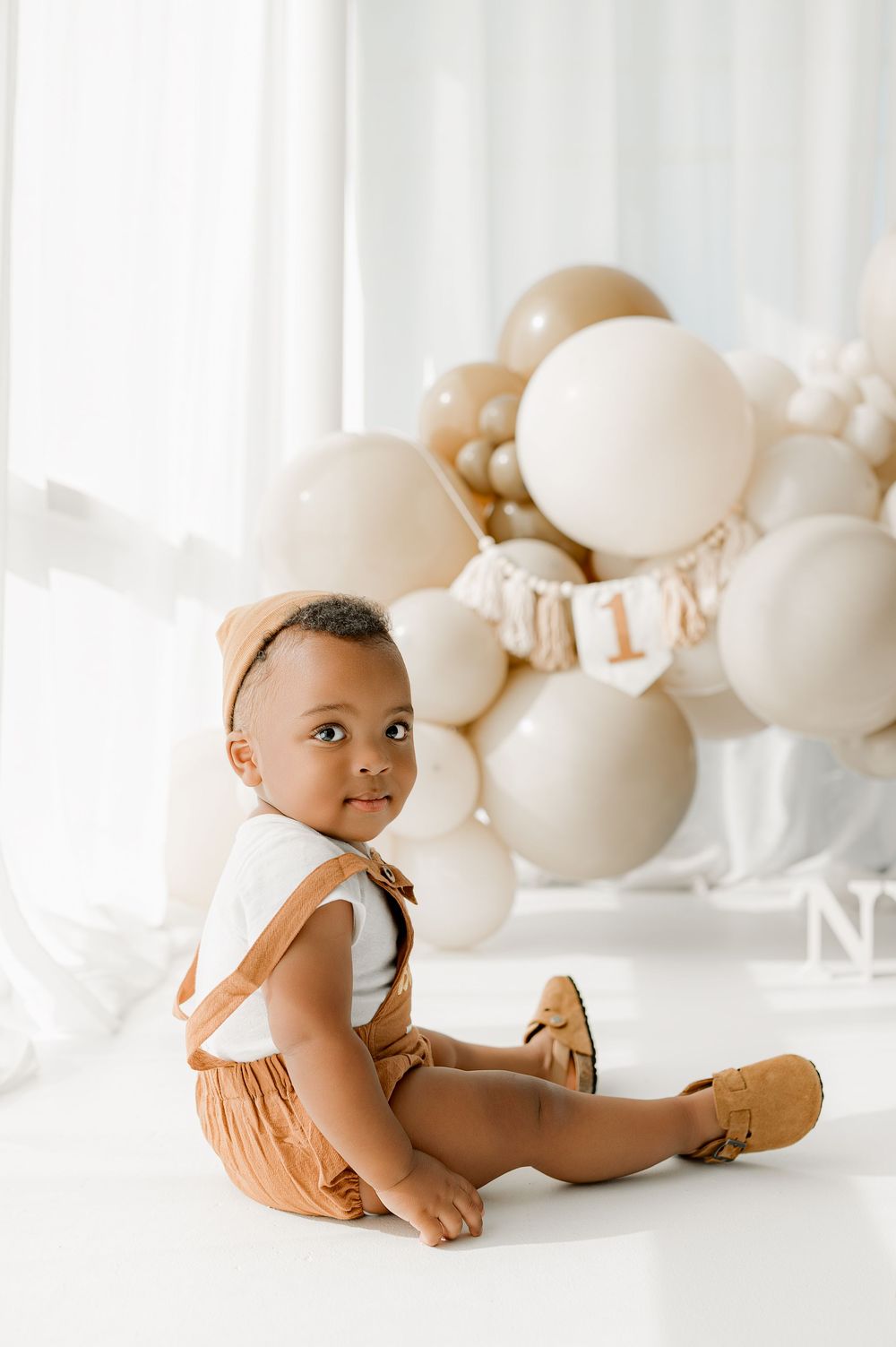 Baby in brown romper sits on white floor with neutral balloon garland decoration in background.