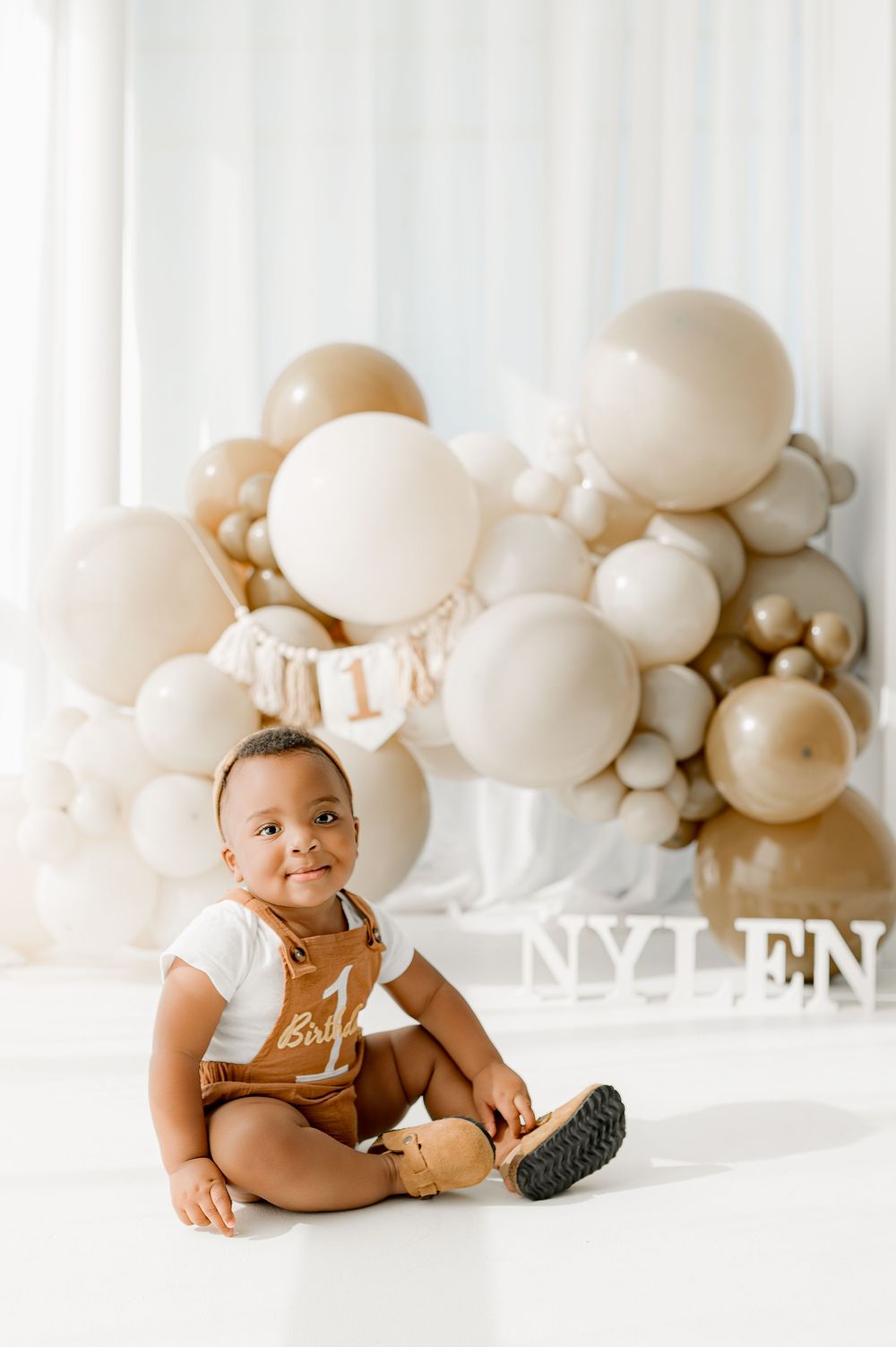 Baby wearing brown romper sitting on white floor with neutral beige balloon arch backdrop in studio setting.
