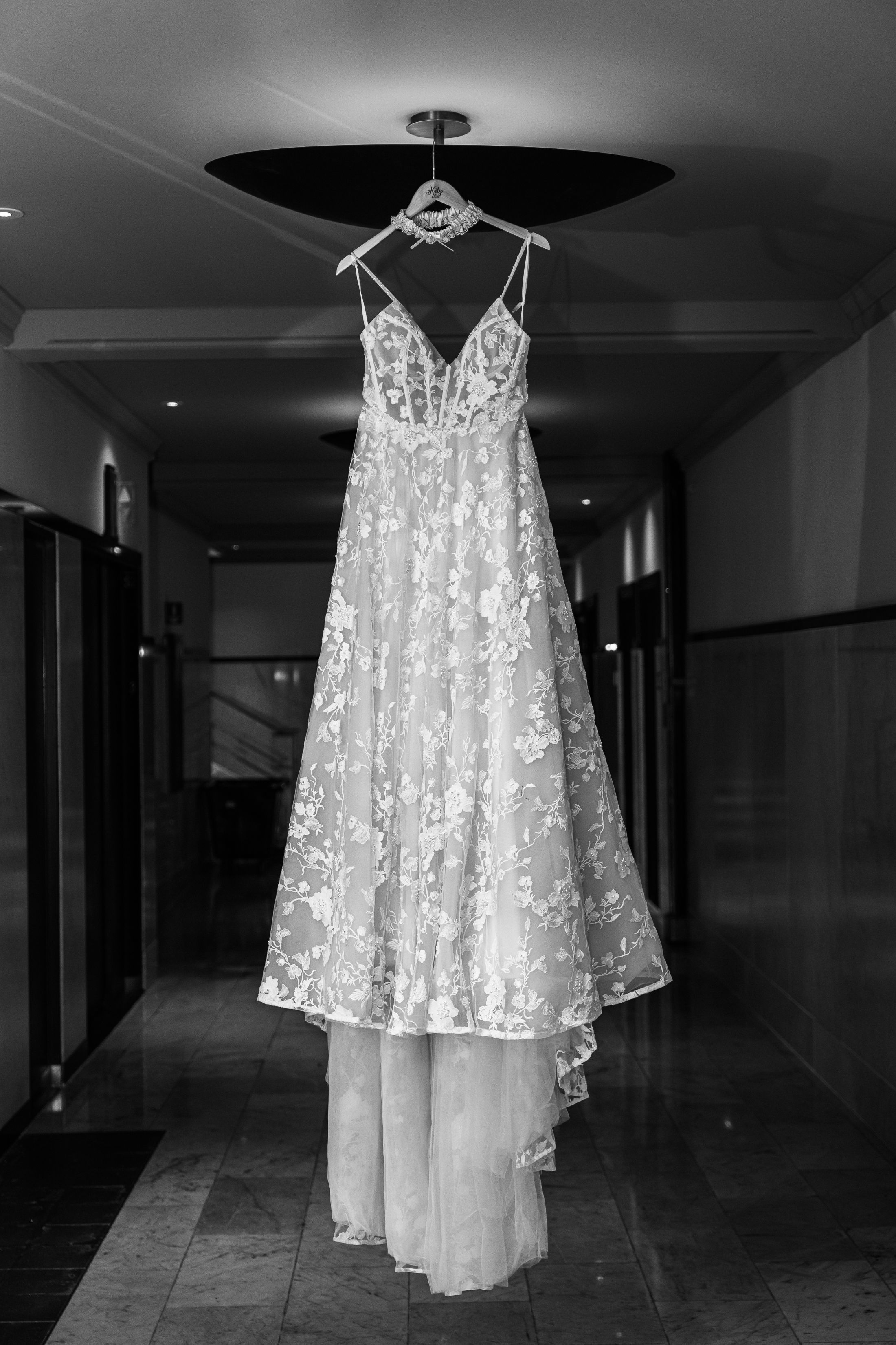 A delicate white lace wedding dress hangs in a dimly lit hotel hallway, showcasing its intricate floral details and flowing design.