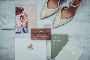 Wedding stationery flatlay with elegant invitations, white pointed bridal shoes and romantic photo on marble surface.
