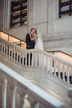 Couple sharing an intimate moment on an elegant marble staircase with white stone balustrades in a grand interior setting.