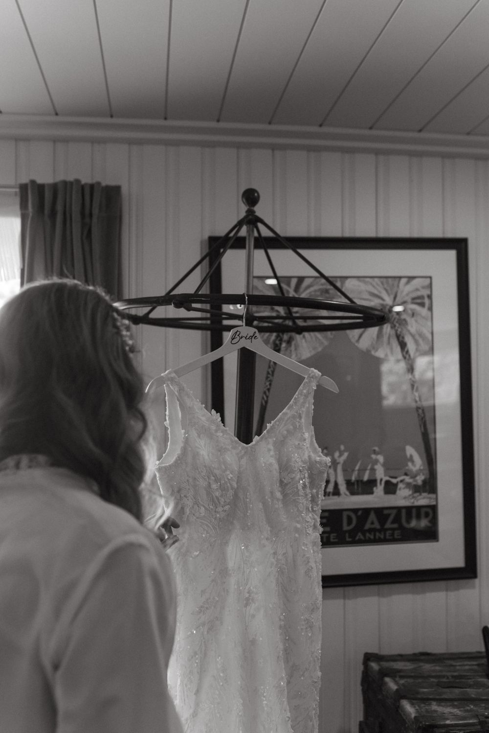 A white lace wedding dress hangs on display in a room with black and white artwork on the walls.