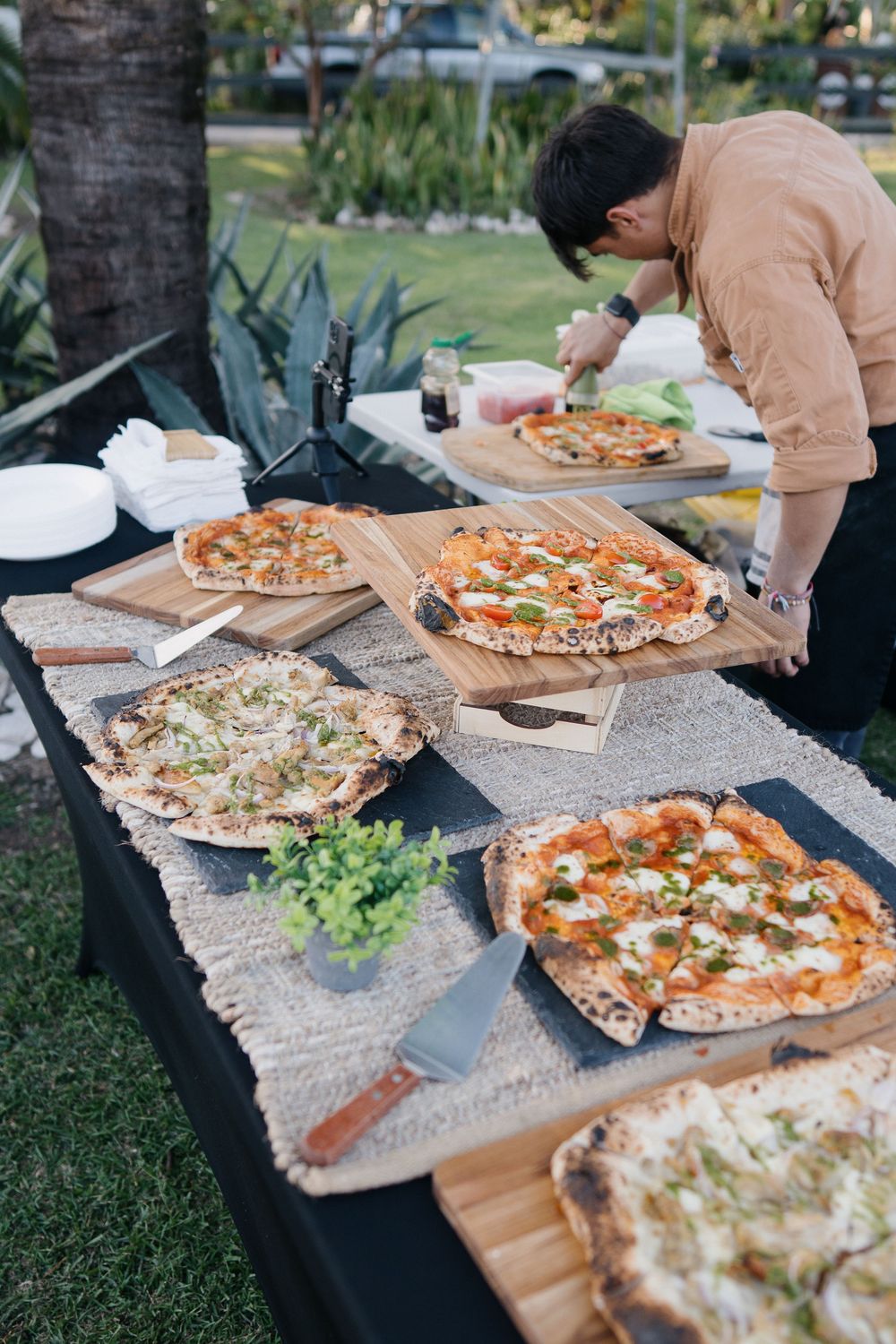 Multiple pizzas with various toppings are laid out on a rustic wooden table outdoors for casual dining.