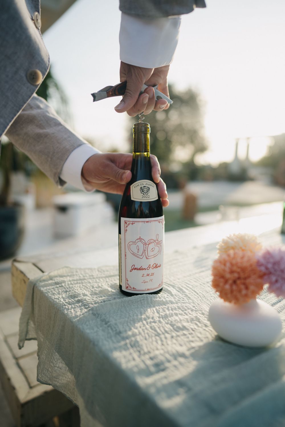 Wine bottle sits on a textured white surface with a soft pink floral arrangement in the background at sunset.