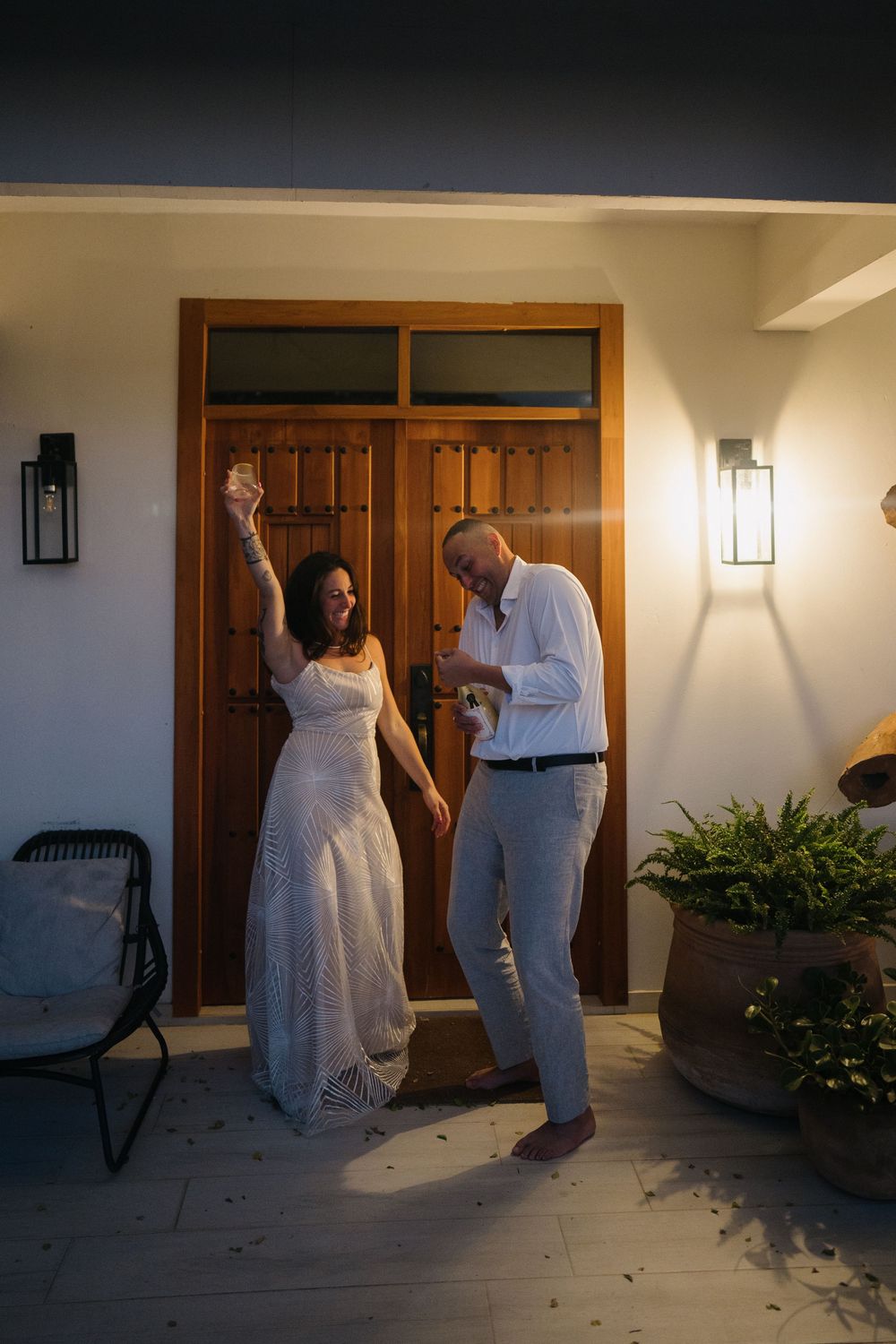 A couple dances together on a porch at sunset with wooden doors and wall sconces in the background.