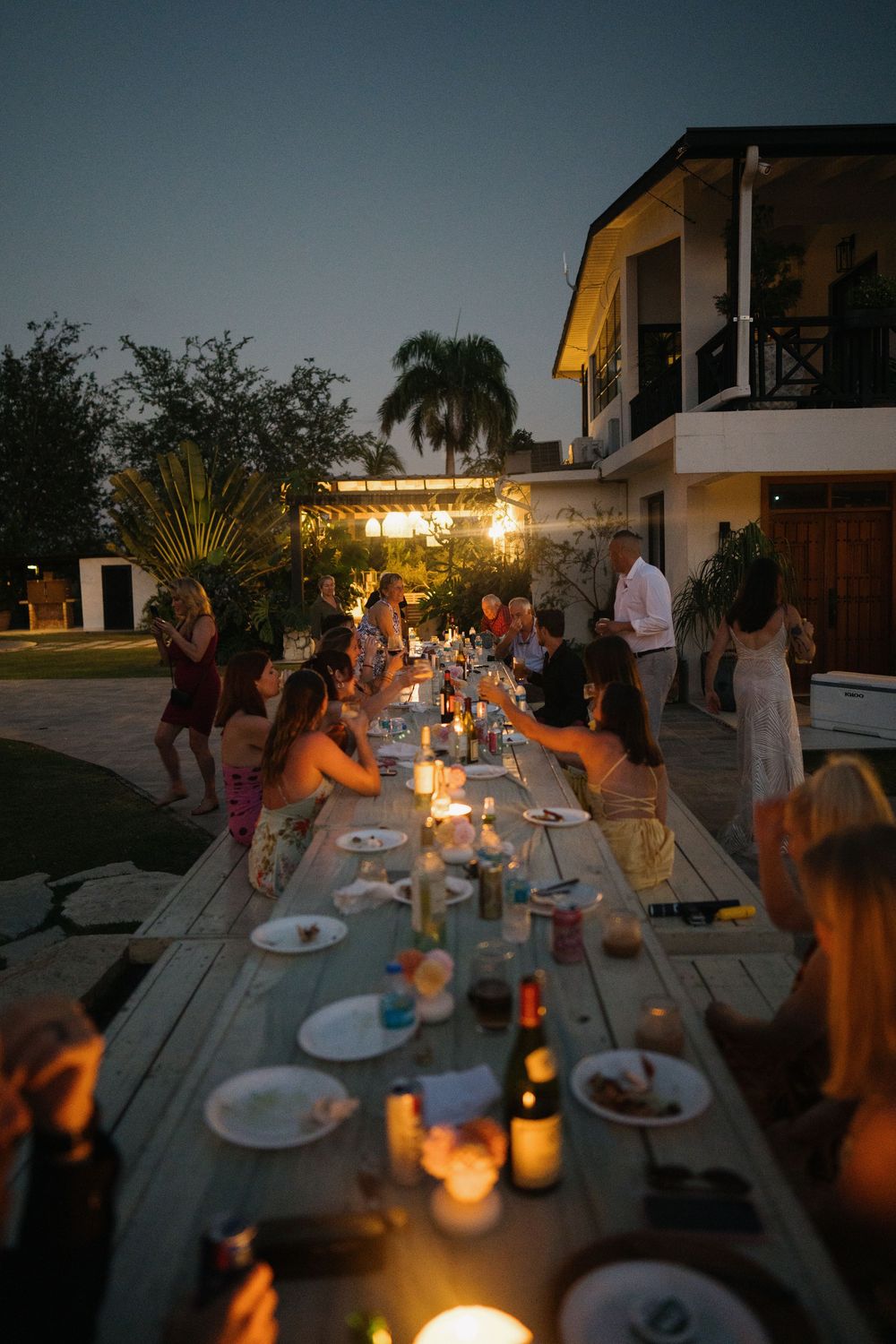 A candlelit outdoor dinner party at dusk with guests seated at a long wooden table under string lights.