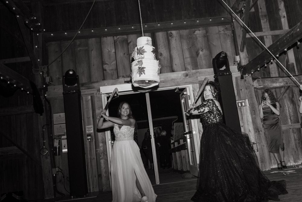 A black and white wedding reception photo showing people dancing in a rustic barn setting with string lights and a tiered cake.