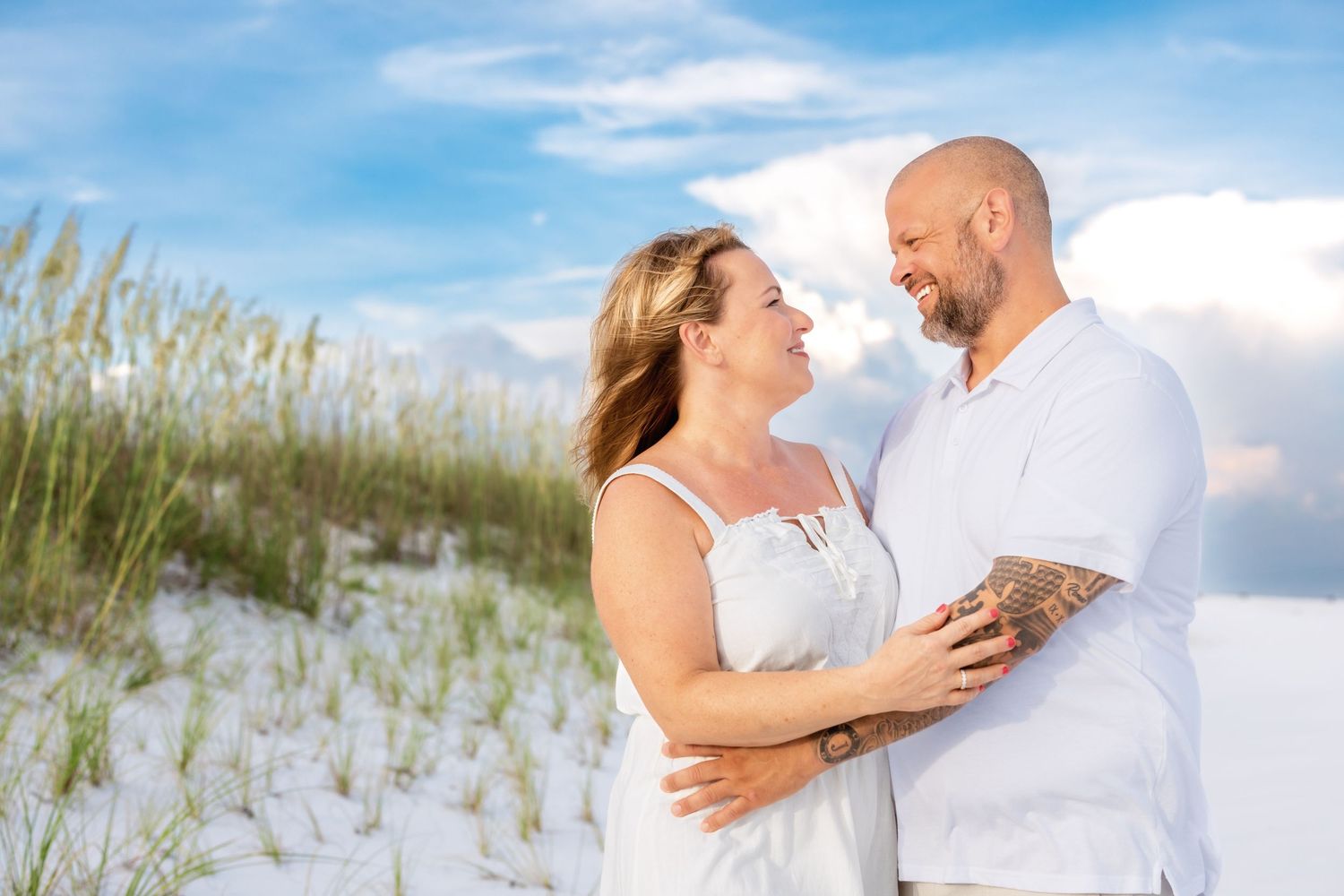 A couple in white outfits shares romantic moments on a sandy beach with sea grass and blue skies in the background.