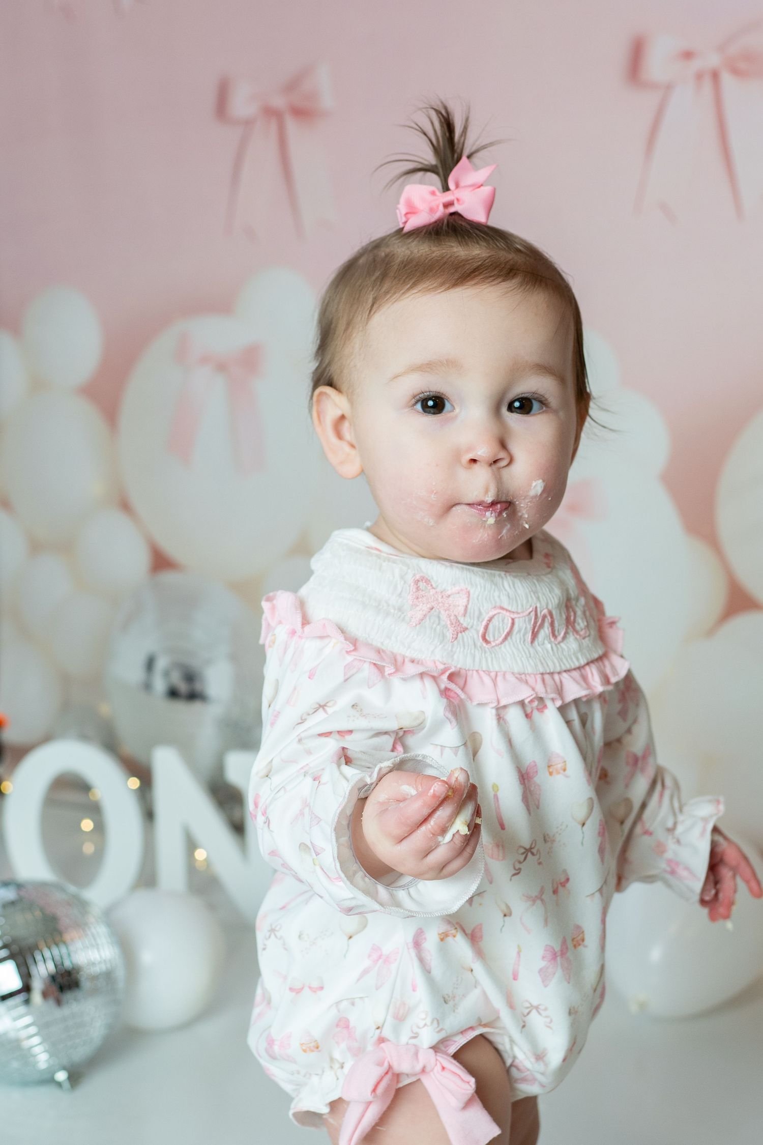 Baby in white and pink floral dress with bow against pastel backdrop with balloons and decorative bows.