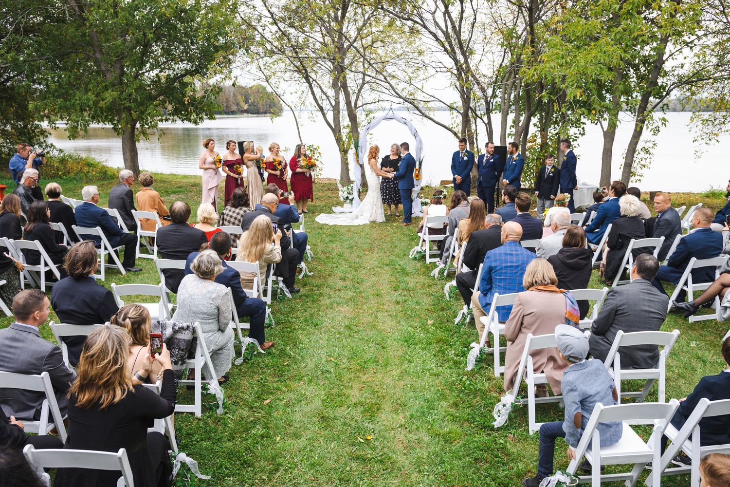 Guests seated in white chairs at an outdoor waterfront wedding ceremony surrounded by lush green trees.