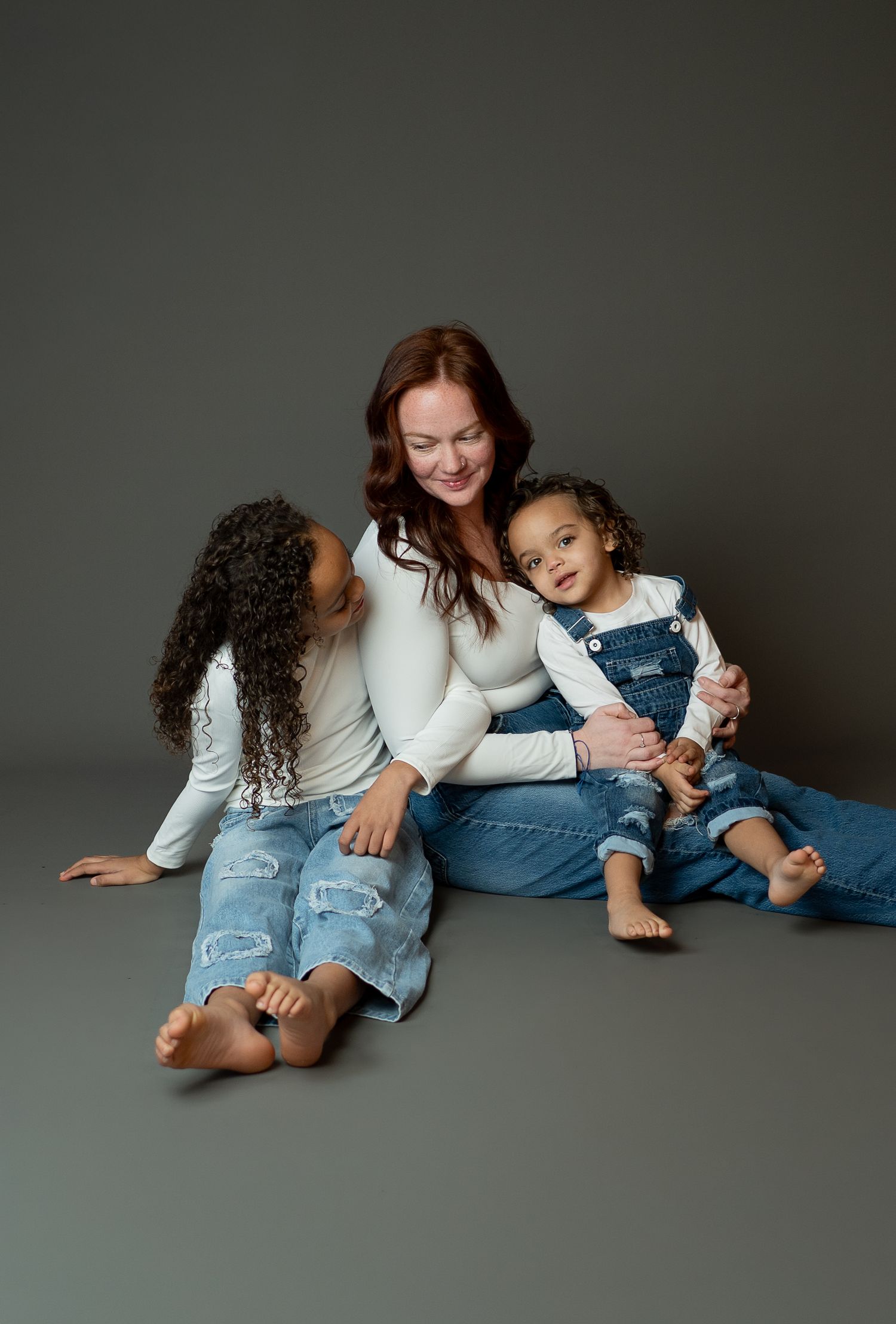 A family wearing white tops and denim sits together on a gray floor, sharing joyful moments in a studio setting.