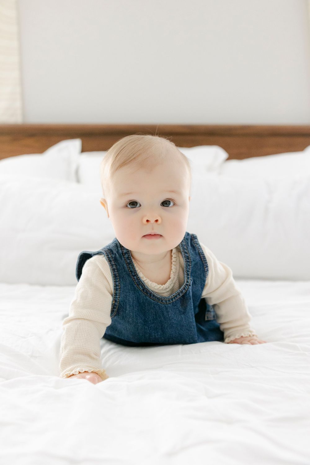 Baby wearing navy blue vest and cream sweater lies on white bedding in soft natural light.