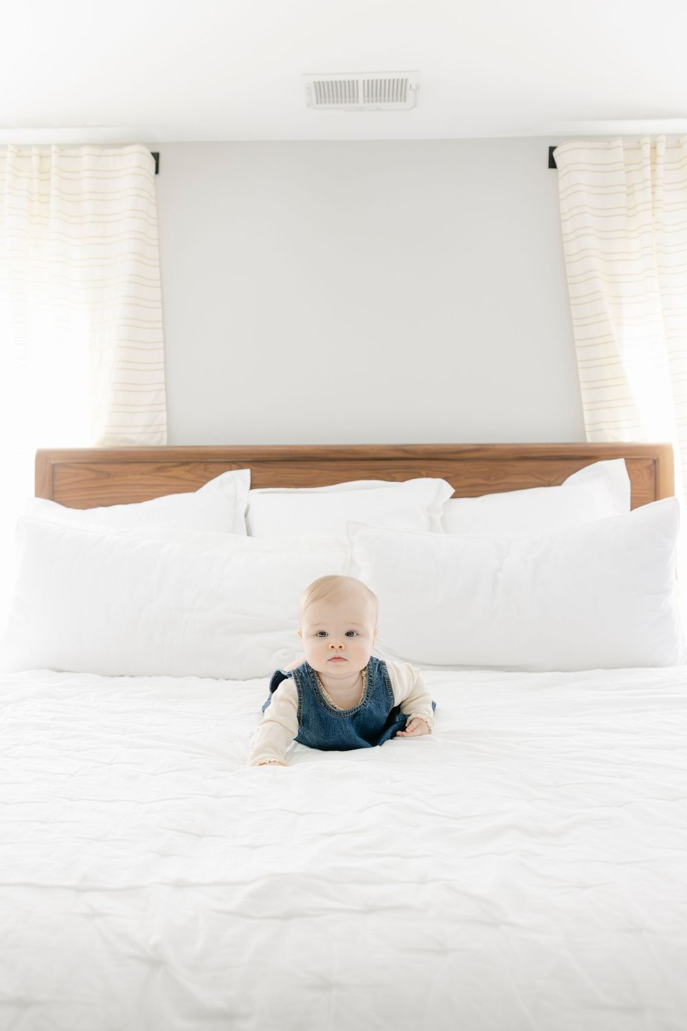 A small baby in a blue jumper lies on a white bed with a wooden headboard and light curtains in the background.
