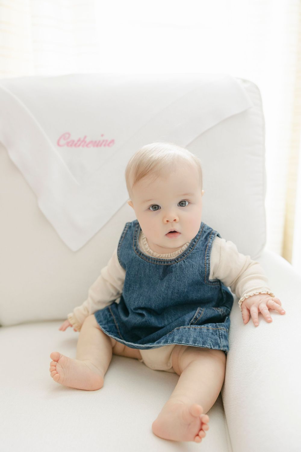 Baby wearing denim dress sits on white couch in series of candid lifestyle portraits.