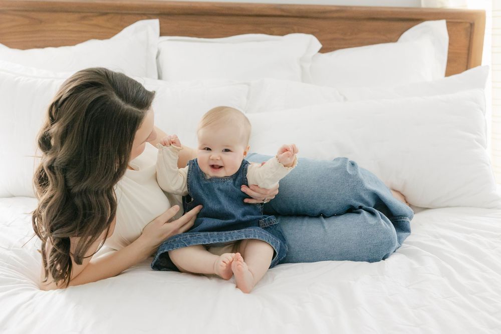 A parent and baby share a tender moment while relaxing together on a white bed with wooden headboard.