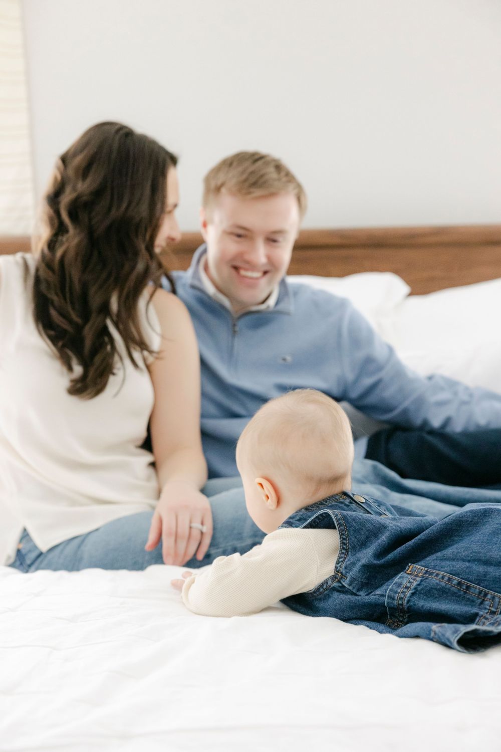 A family lounges together on white bedding wearing casual clothing in a bright, airy setting.