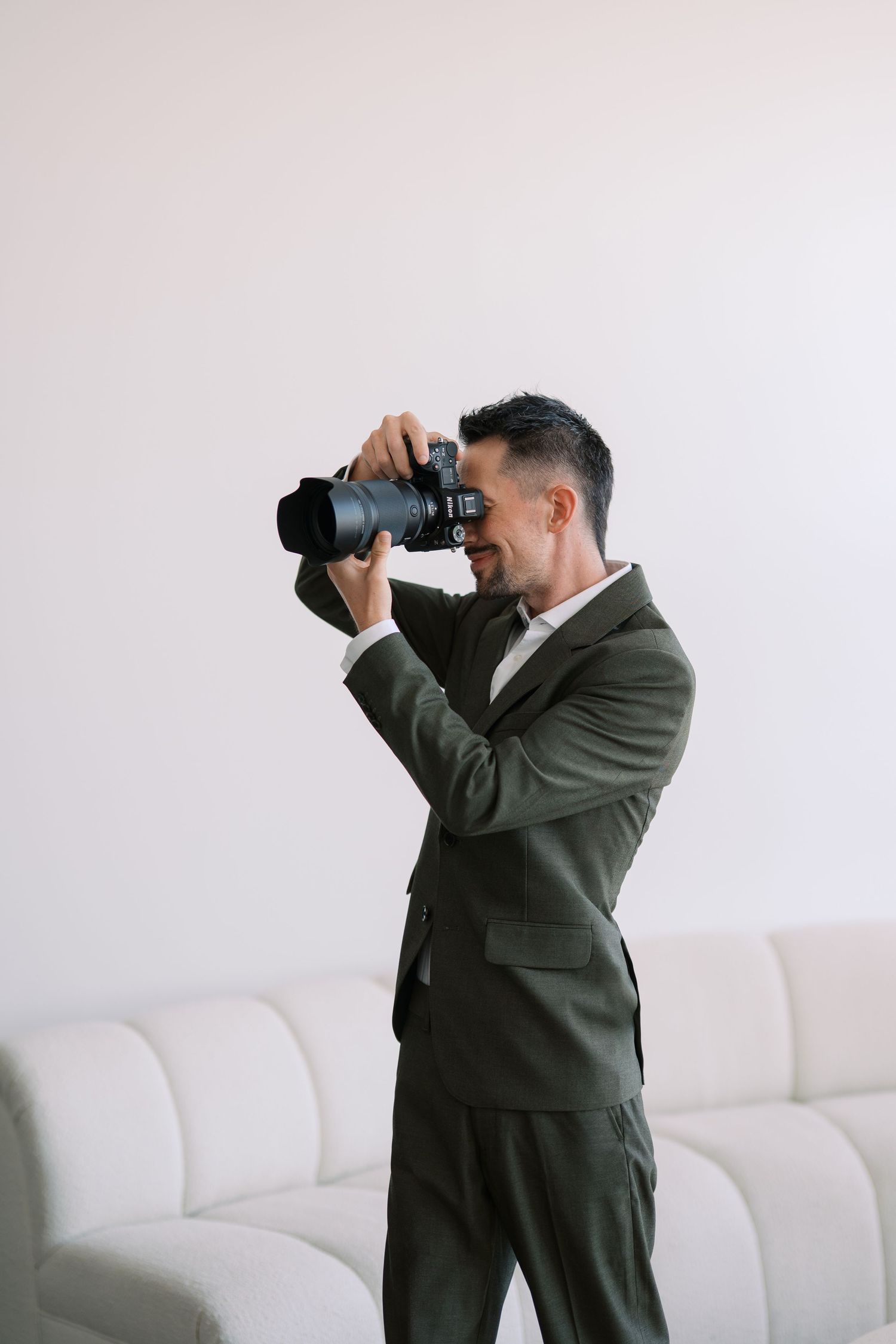 Professional photographer in olive suit taking photos with telephoto lens against white backdrop.