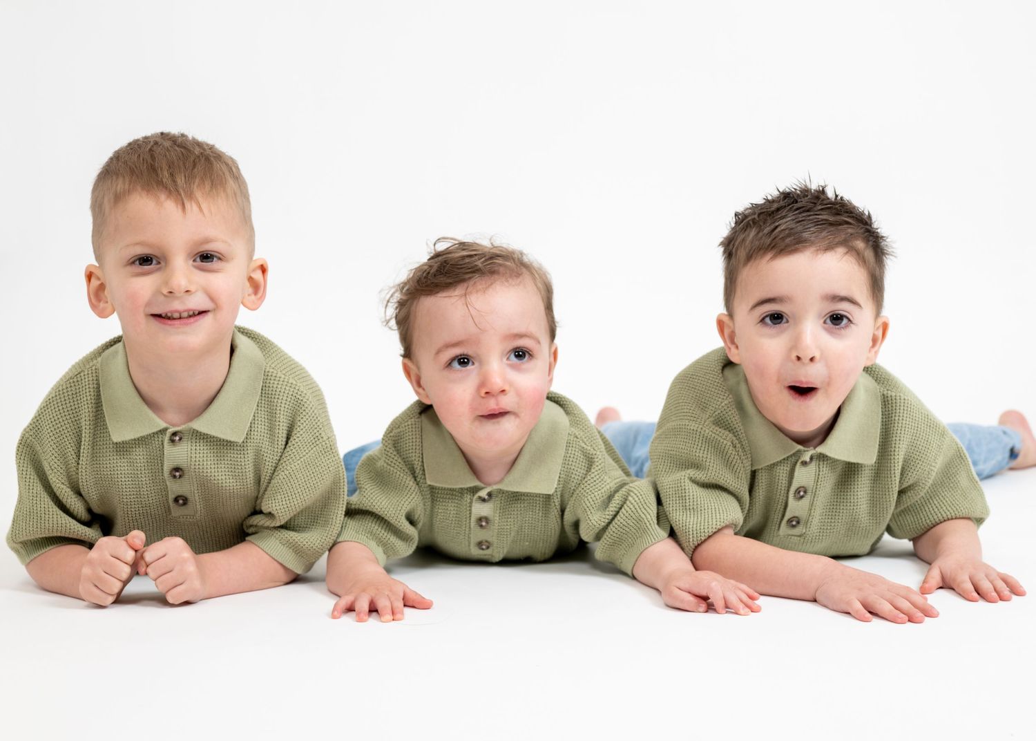 Three young children wearing matching olive green polo shirts lie on their stomachs on a white background.