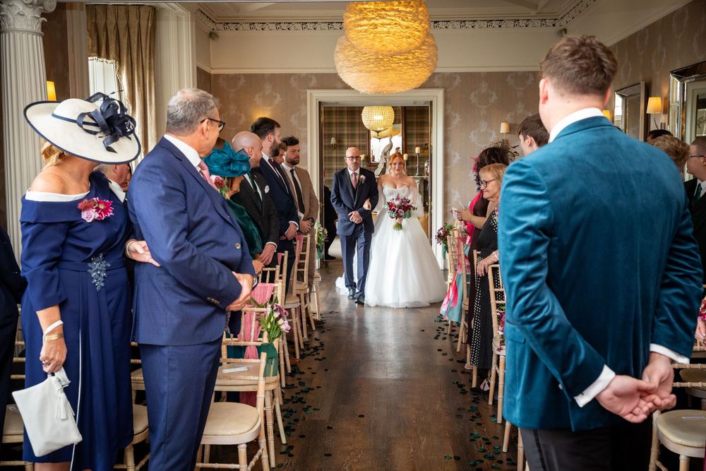 Bride walks down the aisle escorted by family in a bright elegant ceremony room.
