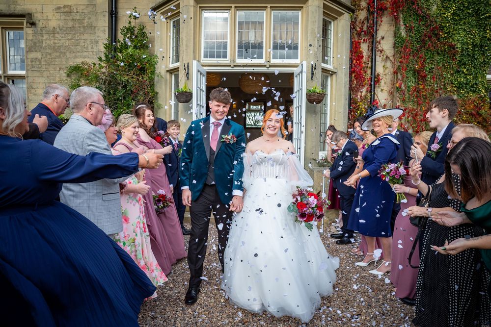 Newlyweds walk through confetti shower outside historic ivy-covered wedding venue with cheering guests.