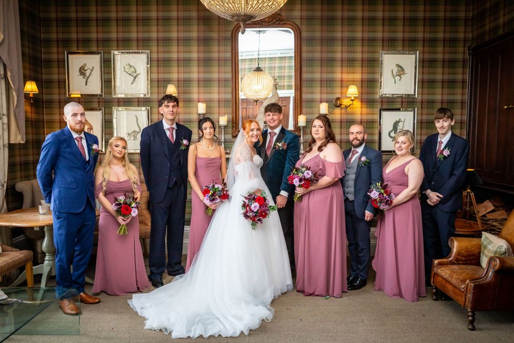 Wedding party poses in tartan-walled room, bridesmaids in dusty rose gowns with red bouquets.