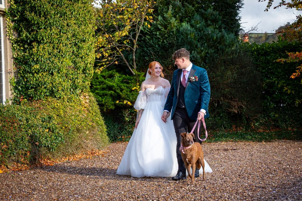 Bride and groom walk dog along gravel path at autumn wedding venue.