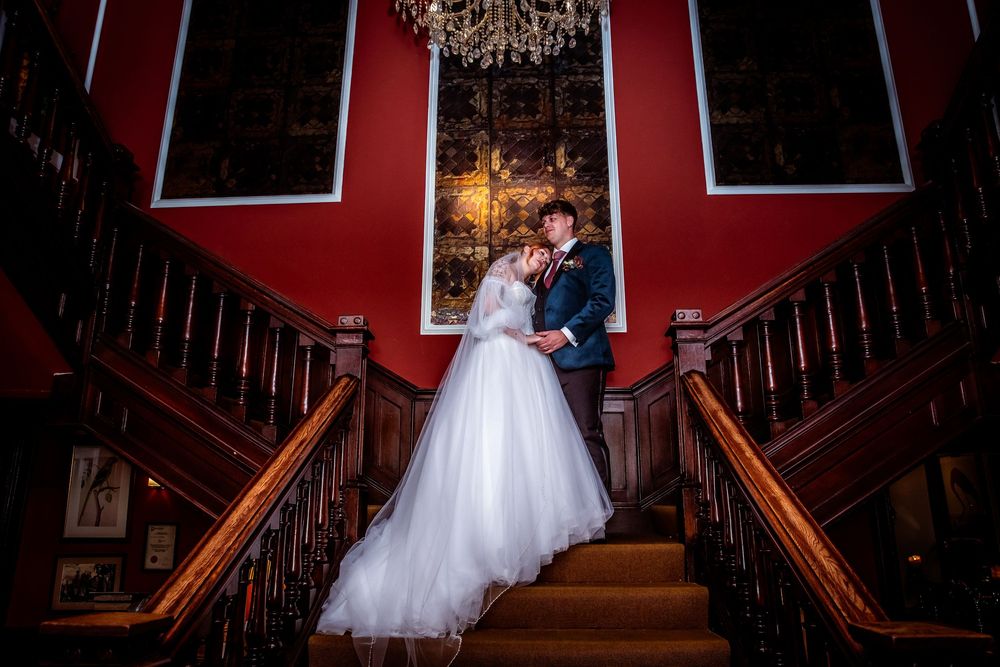 Bride and groom embrace romantically on grand wooden staircase beneath ornate chandelier in red room.