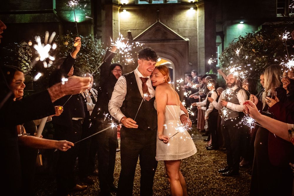 Newlyweds kiss surrounded by sparkler send-off at night outside wedding venue.