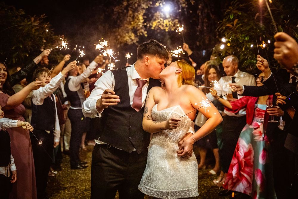 Couple shares romantic kiss during sparkler exit with wedding guests celebrating at night.
